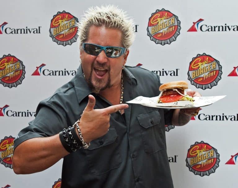 A person with spiky blond hair wearing sunglasses and a dark shirt is pointing at a burger with a large lettuce leaf and tomato. The backdrop features logos of "Guy's Burger Joint" and "Carnival.