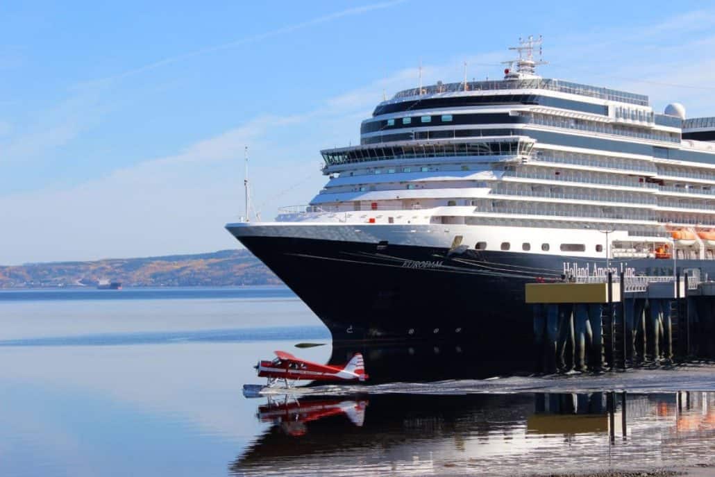 A large cruise ship majestically rests at the port, while a small red seaplane gracefully takes off or lands on the serene water. The sky is clear and blue, with picturesque hills providing a stunning backdrop.