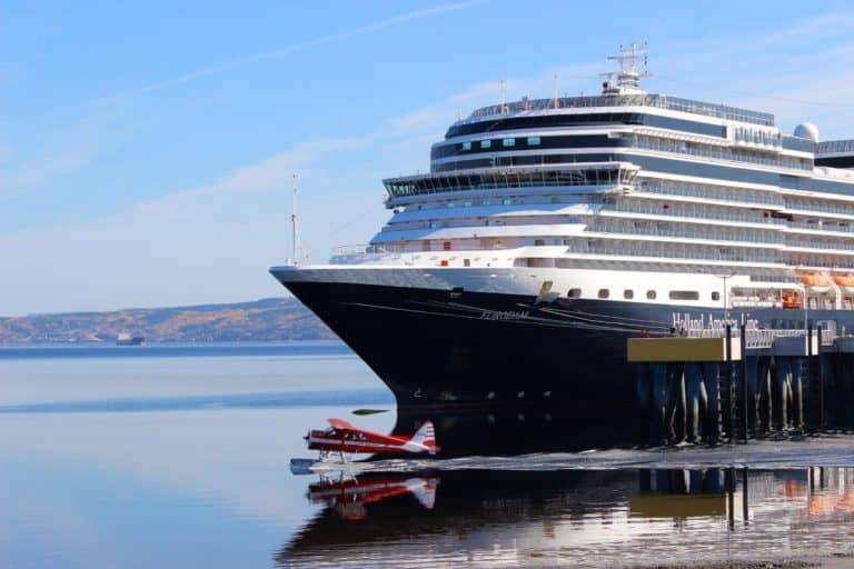 A large cruise ship majestically rests at the port, while a small red seaplane gracefully takes off or lands on the serene water. The sky is clear and blue, with picturesque hills providing a stunning backdrop.