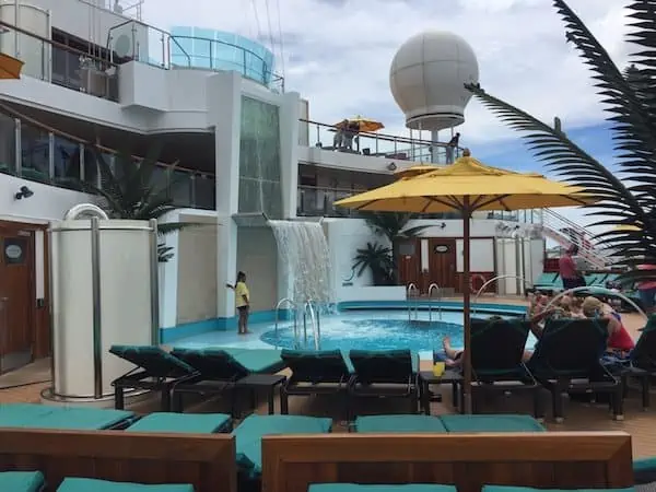 The cruise ship deck boasts a small pool, lounge chairs, and a waterfall feature. Passengers unwind under yellow umbrellas as the large white radar dome stands out against a partly cloudy sky, with lush green plants completing the scene.