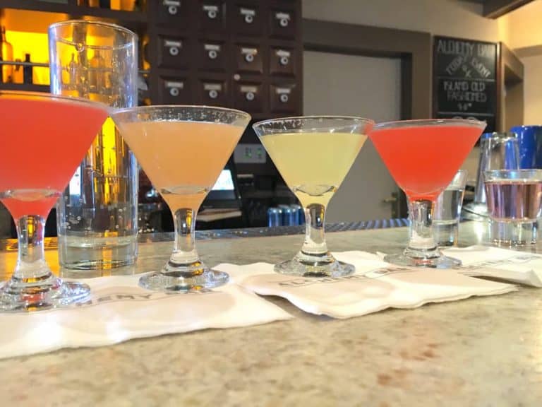 A row of four colorful cocktails on the alchemy bar counter, each in a martini glass. Each drink appears to be a different color, ranging from red to pink to yellow. A tall empty glass and small shot glass are placed next to them. The background features bar shelves.