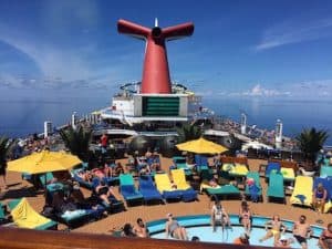 On a clear day, numerous individuals relax poolside on the deck of a cruise ship. The deck features lounge chairs, some with yellow umbrellas, and a pool in the foreground. A red and white funnel is prominently visible against a blue sky—ever wondered how much money to take on a cruise?