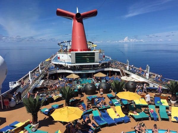A busy cruise ship deck on the Carnival Sunshine features numerous people lounging on colorful chairs and sunbathing. The upper deck boasts a sizable screen, a red funnel, and several palm trees. The ocean and a clear blue sky are visible in the background.