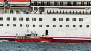 A small orange river pilot boat sails alongside a large white cruise ship. The cruise ship features numerous windows and lifeboats, with one person from the pilot boat visible near an open door on the ship.