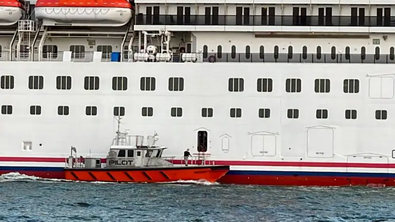A small orange river pilot boat sails alongside a large white cruise ship. The cruise ship features numerous windows and lifeboats, with one person from the pilot boat visible near an open door on the ship.