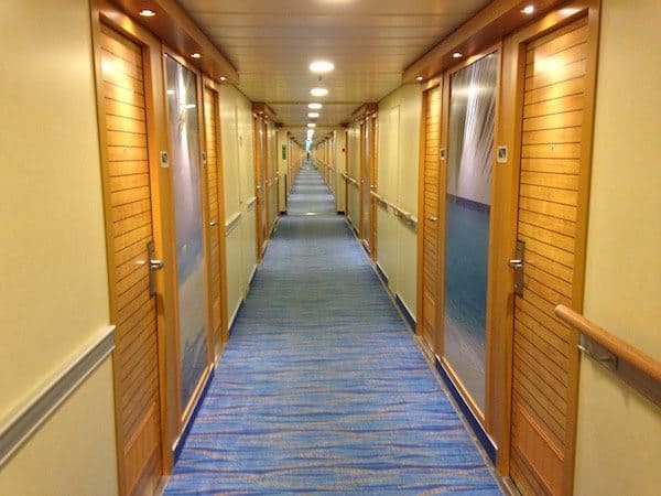 A long hallway on the cruise ship features wooden doors on both sides, each with a metal handle and room number plaque. The carpeted floor, adorned with a blue pattern, complements the light yellow-painted walls. Recessed lighting in the ceiling ensures a bright and welcoming ambiance.