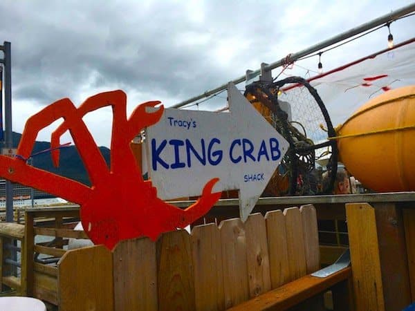 A sign shaped like a red crab with an arrow points towards Tracy's Crab Shack. The sign is mounted on a wooden fence. Various nautical items are visible in the background, including ropes and a large buoy. The sky appears overcast.