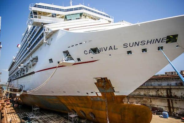 A large white cruise ship named Carnival Sunshine is docked in a shipyard. The hull shows signs of rust and repairs are being performed. The ship's name is clearly visible on the bow, promising future adventures at sea. The background shows a clear blue sky, perfect for cruising.
