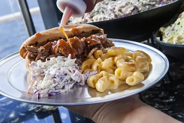A hand holding a metal plate containing a pulled pork sandwich drizzled with sauce, a serving of macaroni and cheese, and a portion of coleslaw. Another hand is squeezing extra sauce onto the sandwich. In the background are bowls of additional food, reminiscent of Carnival Cruise Line BBQ delights.