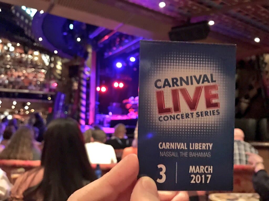 A person holds up a blue Carnival Live Concert Series brochure inside a vibrant venue. The leaflet highlights "Carnival Liberty," "Nassau, The Bahamas," and "3 March 2017." In the background, blurred audience members and a stage illuminated with purple and blue lights add to the excitement.