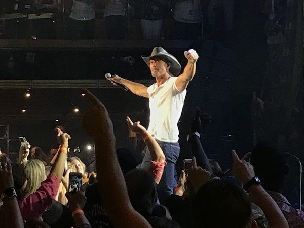 A man wearing a cowboy hat and a white t-shirt is performing live on stage, holding a microphone. He is gesturing with his arms extended outward. The audience below is cheering and pointing towards him with raised hands, giving the scene a carnival-like atmosphere in the dimly lit concert venue.