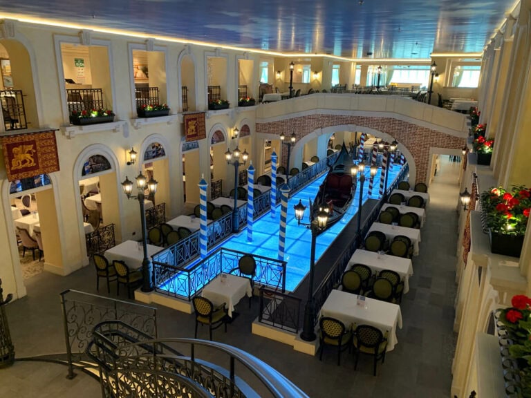 A restaurant with an indoor Venetian-style canal and gondola, surrounded by tables and chairs in the main dining room. The decor includes lanterns, flags, and flowers on railings, all beneath a painted sky ceiling.