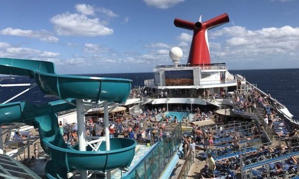 A busy deck on a cruise ship features a teal water slide and numerous sunbathers on lounge chairs. The ship's signature red funnel and a large screen are visible in the background under a partly cloudy sky.