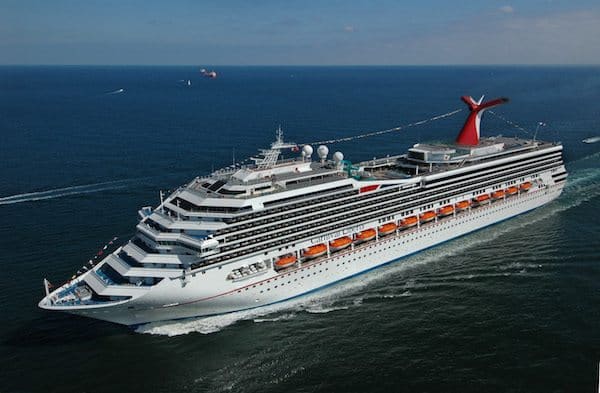 A large white Carnival Cruise ship with a red funnel travels in open water. The ship features multiple decks with balconies and lifeboats. There are a few smaller boats and ships in the background under a clear blue sky.