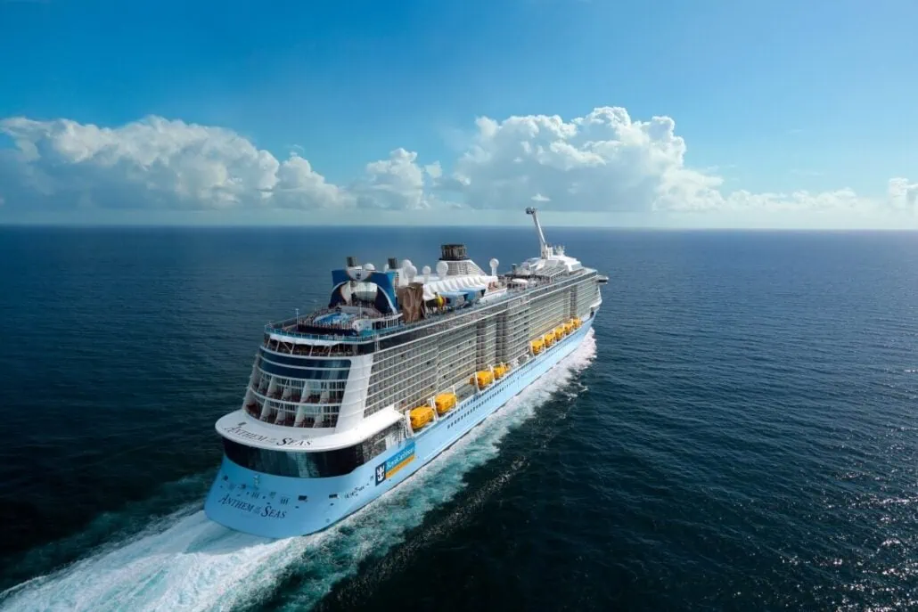 A large cruise ship, Anthem of the Seas, sails through a calm blue ocean on a clear day with a few clouds in the sky.
