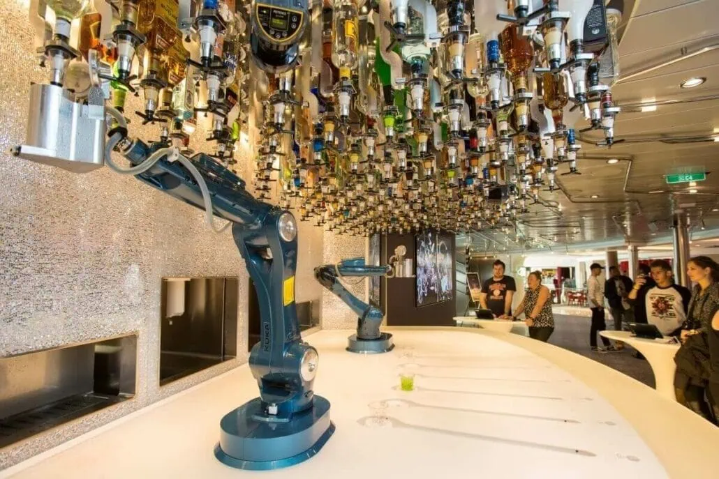 Robotic arms in a bar setting aboard Anthem of the Seas, with numerous bottles hanging upside down from the ceiling, preparing drinks for patrons who are observing in the background.