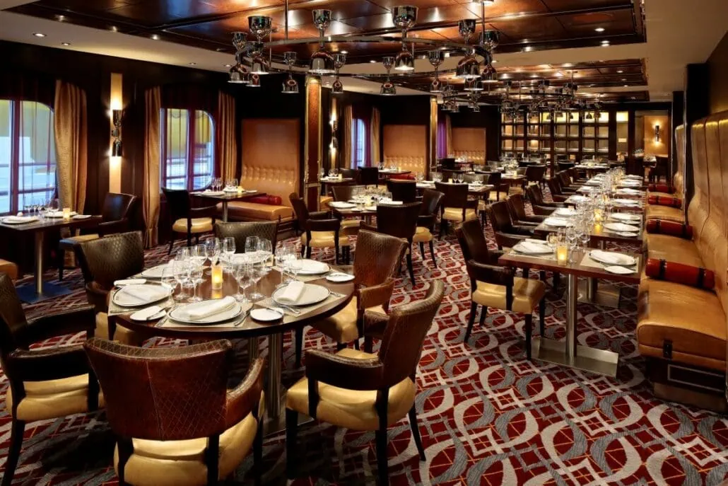 A well-lit, elegant dining room on the Anthem of the Seas with neatly arranged tables set for guests. The room features wooden accents, patterned carpeting, cushioned chairs, and ceiling-mounted lighting fixtures.
