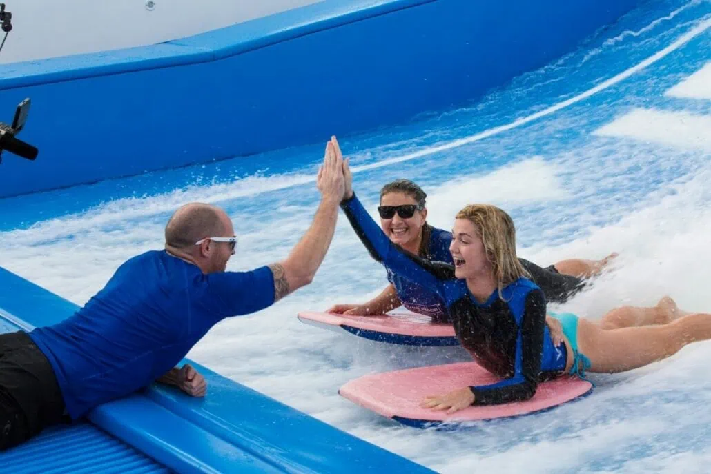 Two people on bodyboards high-five a man in a blue shirt while riding an artificial wave at the surf park aboard Anthem of the Seas.