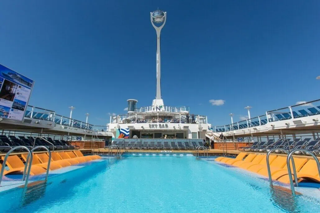 Aboard Anthem of the Seas, the cruise ship deck features a swimming pool with lounge chairs, surrounded by seating areas and the Sky Bar in the background, all under clear blue skies.