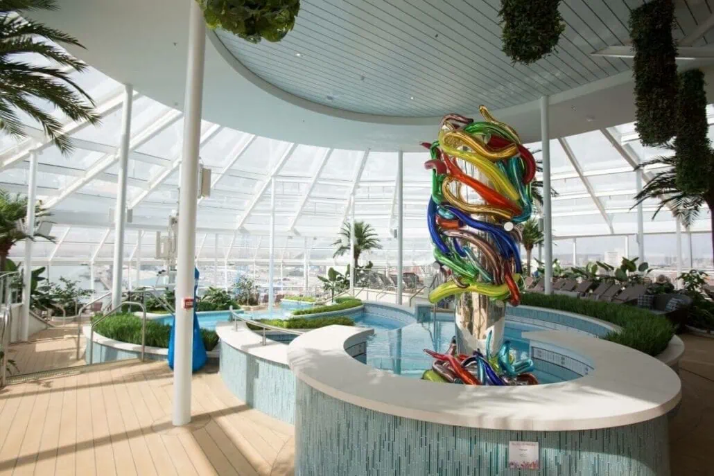 A modern indoor space on the Anthem of the Seas, featuring a colorful abstract sculpture by a circular pool, surrounded by plants and lounge chairs under a glass-paneled roof.