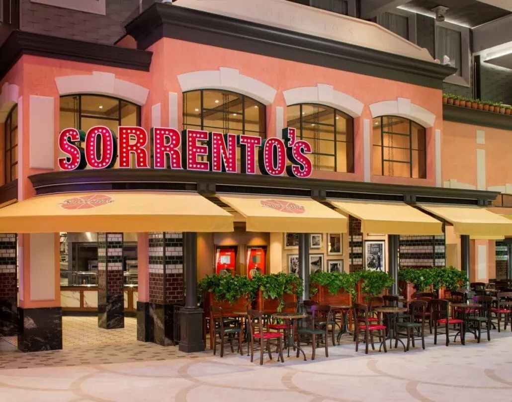 Exterior of Sorrento's pizzeria with red signage, yellow canopies, outdoor seating, and potted plants evokes a charming Mediterranean vibe. Often bustling with visitors from Anthem of the Seas, this spot offers a delightful dining experience under the sun.