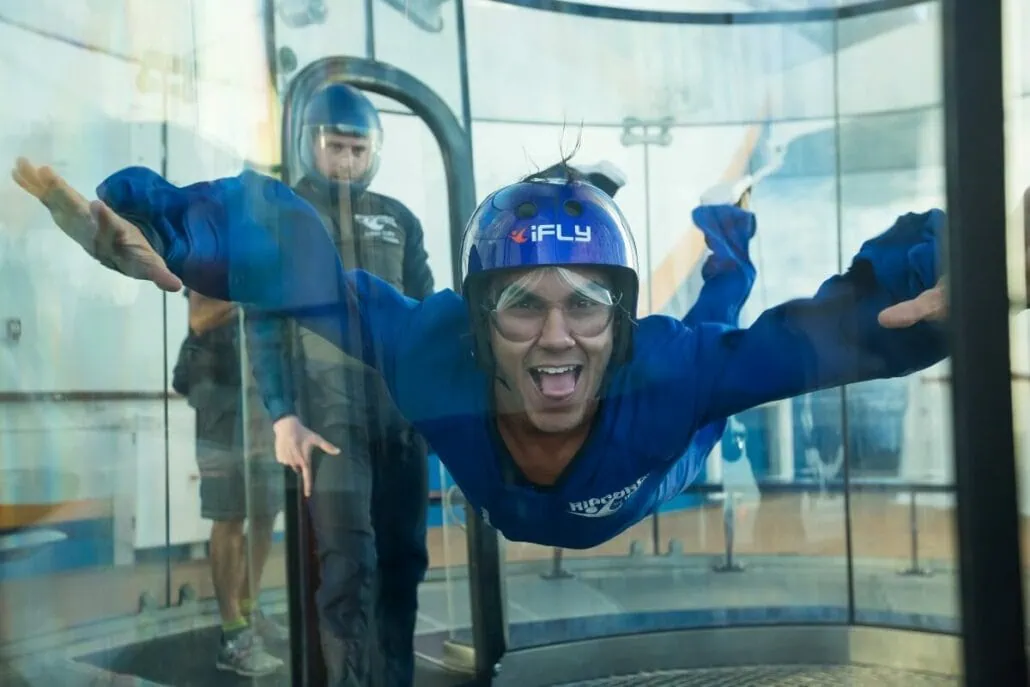 A person in a blue suit and helmet is floating in a vertical wind tunnel, simulating skydiving, aboard the Anthem of the Seas, while another individual stands nearby.