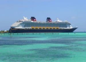 A large white cruise ship with prominent red chimneys and yellow lifeboats, reminiscent of the Disney Fantasy, is anchored in clear blue water. A wooden pier extends from the shore toward the ship under a crisp, clear blue sky.
