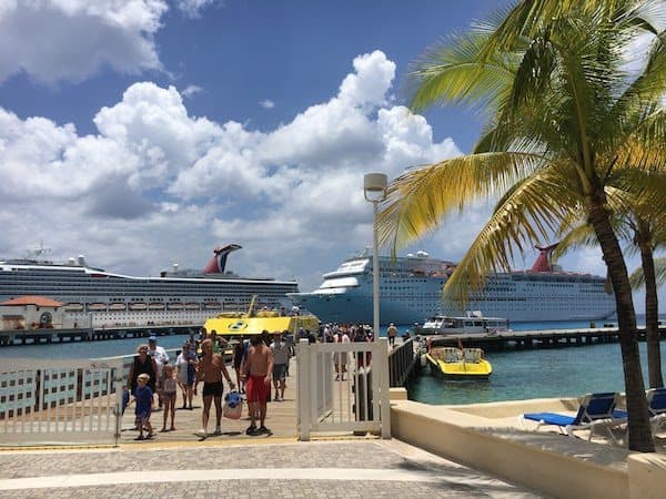 A sunny dock scene in Cozumel with two large cruise ships in the background, people walking on the pier, and a couple of smaller boats nearby. A palm tree is in the right foreground and a few clouds dot the blue sky.