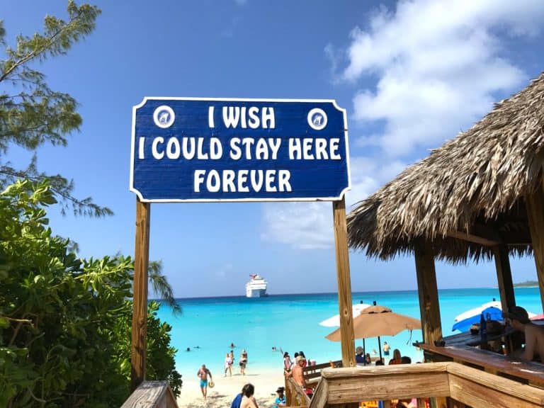 A beach scene with crystal-clear blue water and a blue sky at Half Moon Cay. In the foreground, a wooden sign reads "I WISH I COULD STAY HERE FOREVER." There is a thatched-roof structure to the right, beachgoers with umbrellas, and a cruise ship in the background.