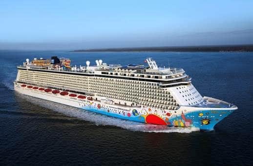 A large Norwegian Breakaway cruise ship travels through calm waters. The ship has multiple decks, numerous windows, and colorful artwork on the front. The sea and a distant coastline are visible in the background under a clear sky.