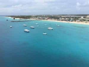 Aerial view of Grand Turk's coastal area with clear blue water and several boats anchored or floating. The shoreline features sandy beaches, numerous buildings, and a Carnival Cruise Line ship that recently returns. The horizon displays a mix of clouds and blue sky.