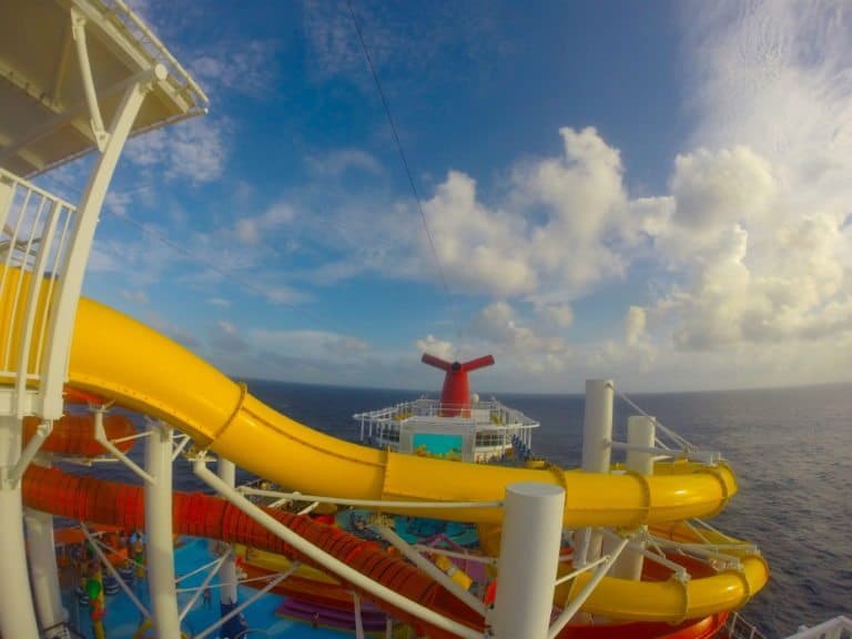 A view of Carnival Vista's upper deck features multiple yellow and red water slides. The ocean is visible in the background under a partly cloudy sky, with the ship's funnel showcasing its distinctive design at the center. This scene captures a moment of fun and relaxation on a trip from Miami.