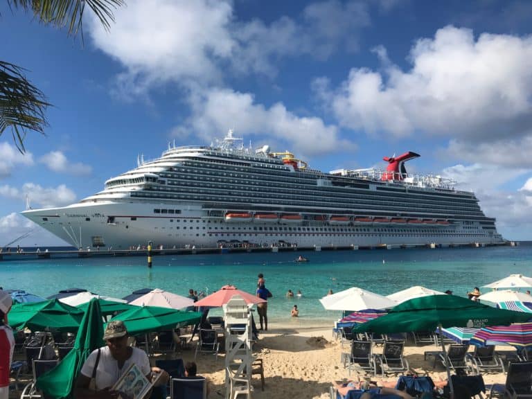 A large cruise ship is docked near Grand Turk beach, with several people visible swimming in the water. The beach is lined with green and white umbrellas and numerous lounge chairs. The sky is mostly clear with a few scattered clouds, while Jack's Shack adds a lively touch to the scene.