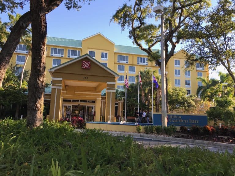Exterior of the Hilton Garden Inn near Fort Lauderdale Airport with a yellow façade and green roofing. The entrance features a portico with the hotel logo above, accompanied by various flags. Trees and plants in the foreground enhance the landscape, making it a convenient stay near the cruise port.