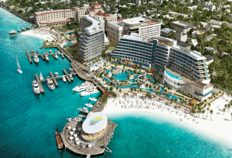 Aerial view of a new resort in the Bahamas featuring several large buildings, multiple pools, and a white sandy beach. There are boats docked at a marina to the left and a stage with a circular roof on a pier in the foreground. The water is a vibrant blue.