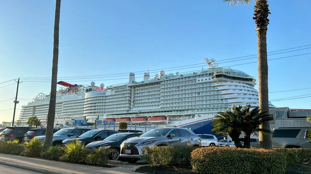 A large cruise ship is docked at the Port of Galveston, visible in the background. Several parked cars and palm trees line the foreground, while a clear blue sky provides a picturesque backdrop for those navigating this vibrant area.