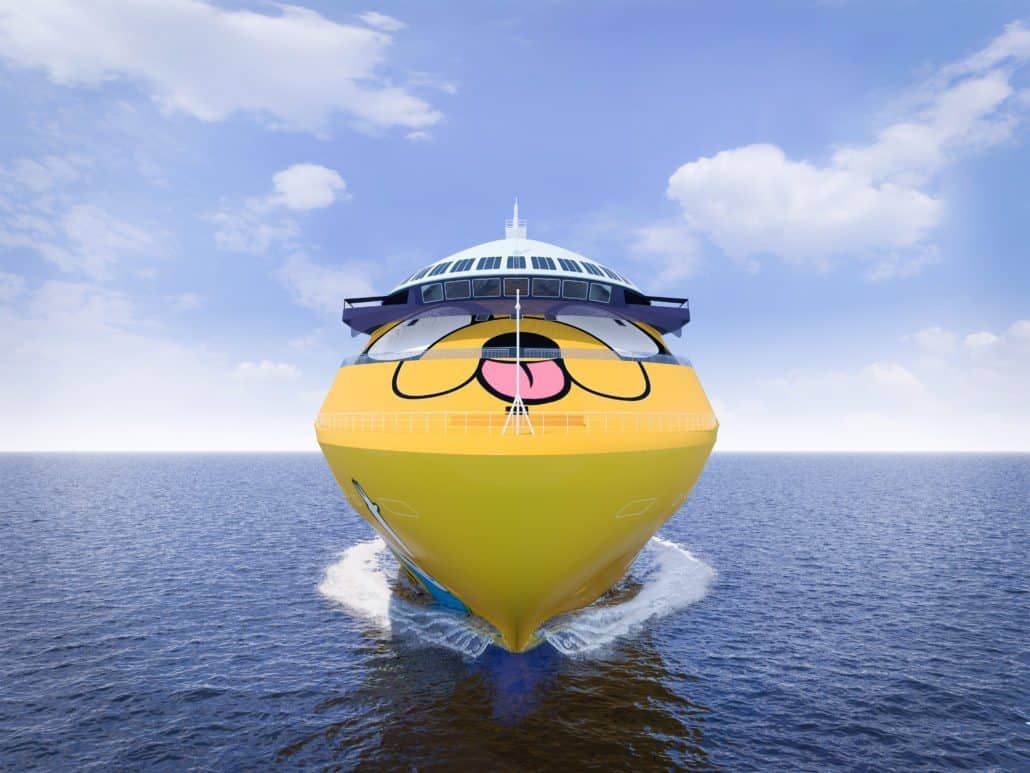 Front view of a yellow cruise ship sailing in the ocean with a cartoon dog's face painted on the bow. The sky is clear with a few clouds, perfect for a family vacation with kids.
