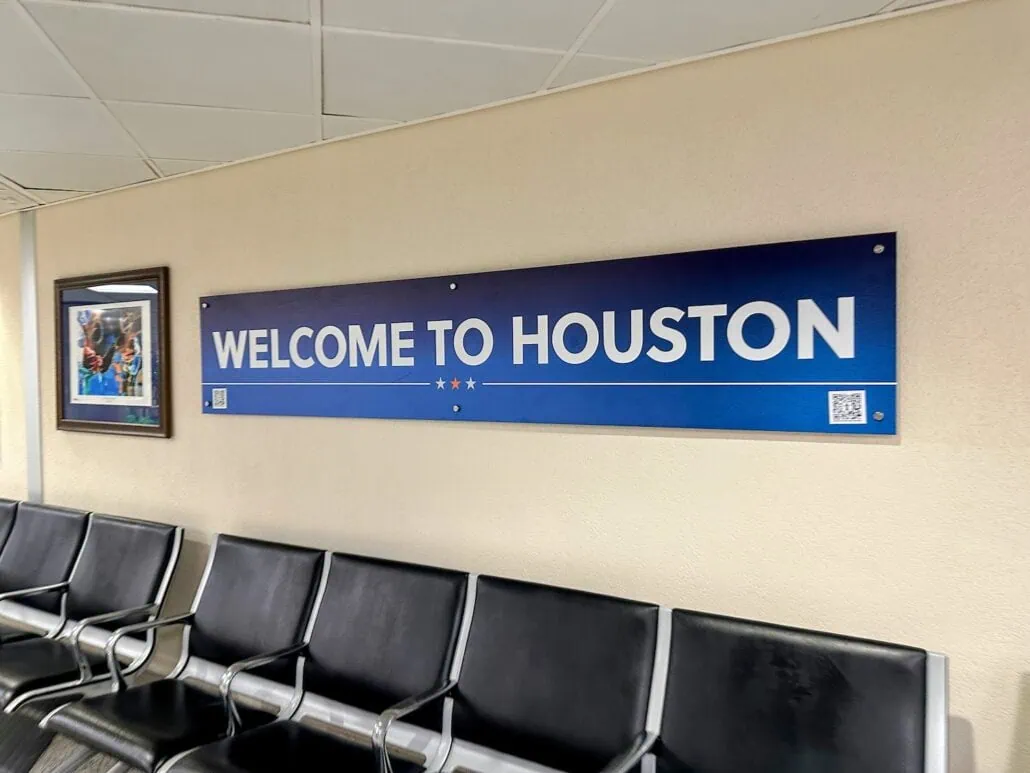 A blue sign on the wall reads "Welcome to Houston" in white capital letters. Below, rows of black chairs are neatly arranged. A framed picture hangs to the left, subtly hinting at nearby attractions like navigating through the bustling Port of Galveston just beyond.