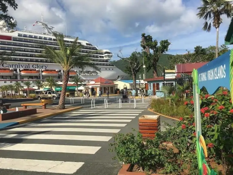 A large cruise ship named Carnival Glory is docked at a port. In the foreground, a zebra-striped pedestrian crossing leads to various buildings and palm trees. A sign to the right partially reads "Caribbean Islands," and there are plants and flowers lining the walkway.