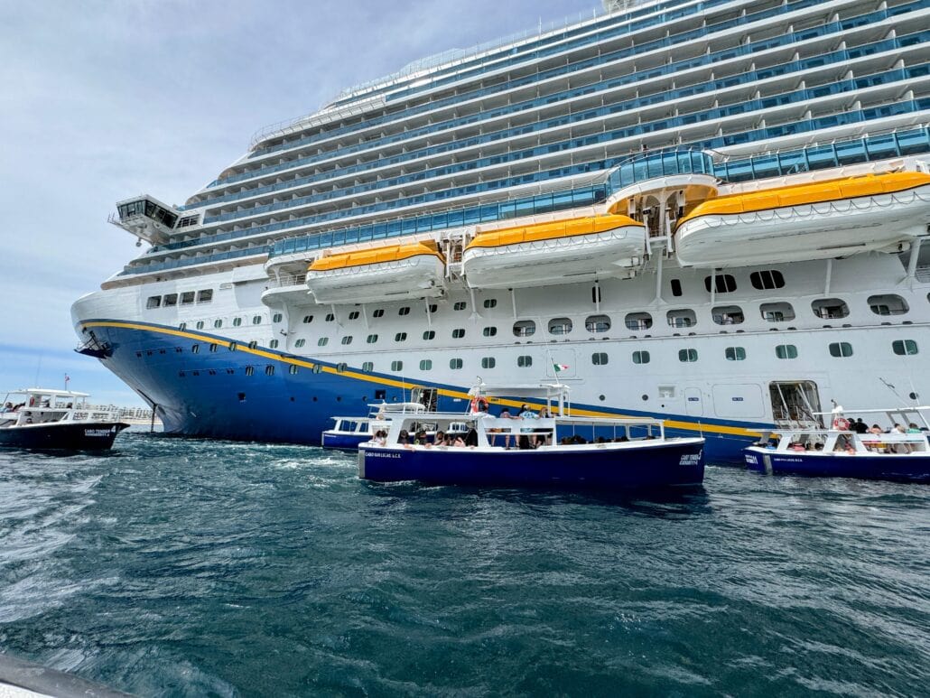 A large cruise ship is docked near the shore, primed for disembarkation, with four small blue and white boats floating alongside it on a sunny day.