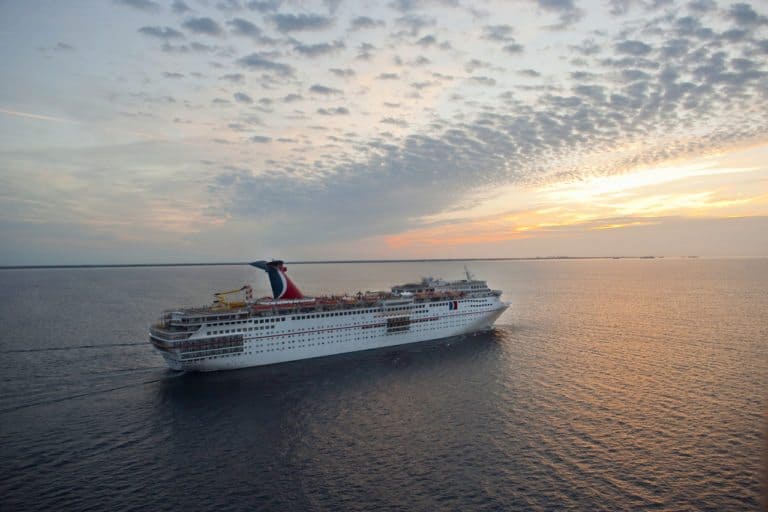 A large cruise ship sails on the open sea under a partly cloudy sky during sunset. The sunlight casts a warm glow on the water, and the ship, reminiscent of a lively Carnival bound for Cuba, is headed towards the horizon.