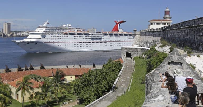 A large cruise ship, Carnival Paradise, sails near an old fort with stone walls. In the foreground, several people observe the ship and fort, with some taking photos. Palm trees and historical buildings are visible, and a lighthouse stands in the distance under a clear sky.
