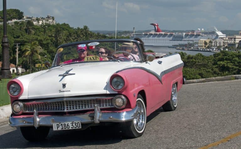 A pink and white vintage convertible taxi with three passengers on a road. In the background, a large cruise ship from Cuba Cruises is docked near lush, green foliage. The scene is set on a sunny day with a clear sky, reminiscent of the vibrant Carnival atmosphere in 2019.