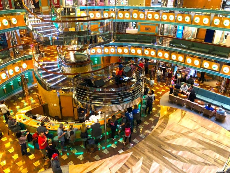 A brightly lit indoor area featuring a bar with people seated and standing around. The space includes multiple levels and a shiny spiral staircase, invoking a touch of Carnival Magic. Walls are decorated with round lights. Several people are visible, engaging in various activities.