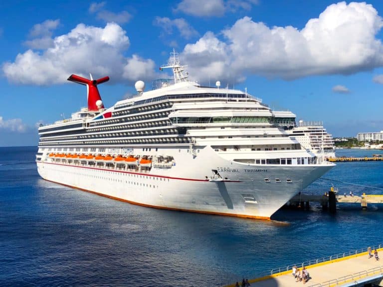 A large white cruise ship named "Carnival Triumph" is docked at a pier on a sunny day with a blue sky and scattered clouds. The ship features multiple decks, balcony doors, lifeboats, and a distinctive red and blue funnel. Surrounding waters are calm and blue.