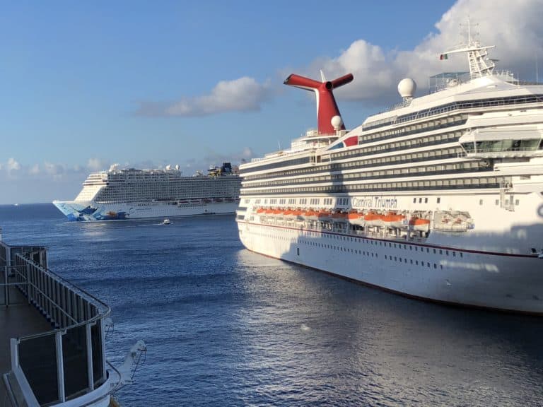 Two large cruise ships are docked side by side in a harbor. The ship in the foreground, named "Carnival Triumph," has multiple lifeboats visible. The background ship, slightly obscured, is likely the "Carnival Magic." The water is calm and the sky is partly cloudy.