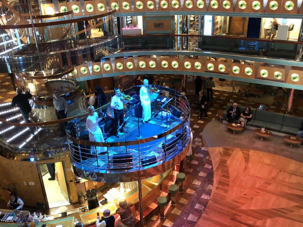 A double-level cruise ship atrium aboard the Carnival Magic features a live band performing on a circular stage. The floor is tiled and wooden with various seating areas. Passengers, including those excited for their Cozumel adventure, gather around, enjoying the music amidst lights and reflective surfaces.