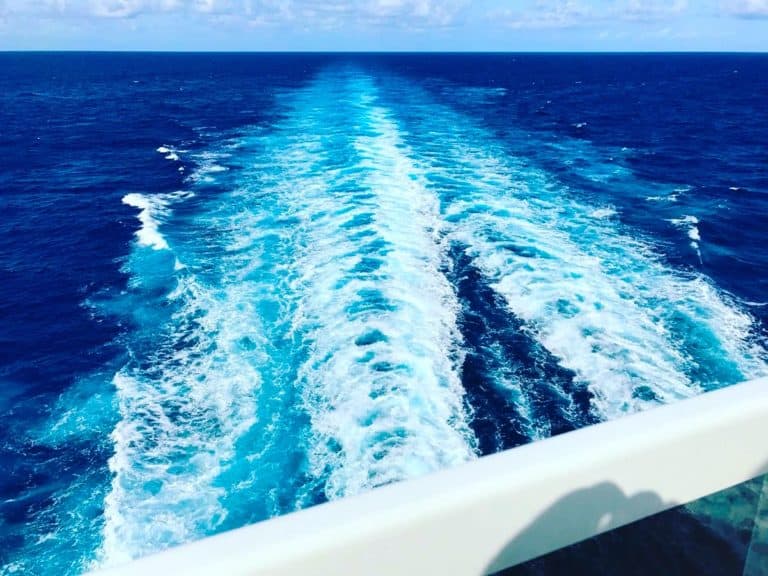 A vast expanse of deep blue ocean with a wide white wake, illustrating the path of a ship moving forward. The horizon is visible with a few scattered clouds in the sky. A white railing is partially visible in the foreground.