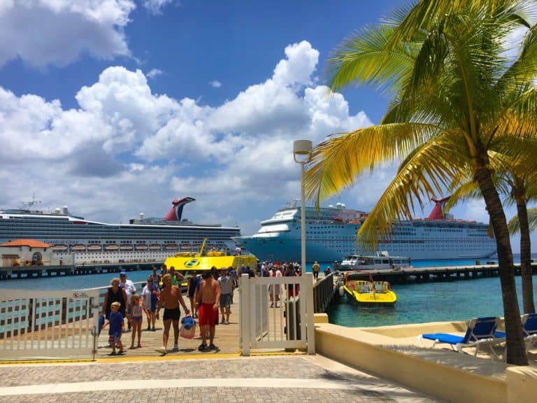 People walking and standing near a dock with two large cruise ships in the background. There are two yellow water vehicles nearby, a palm tree on the right, and partly cloudy skies overhead—a typical scene for those on Mexico cruise excursions.