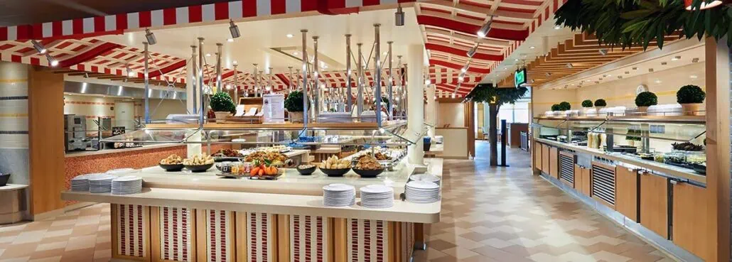 A buffet restaurant reminiscent of a Carnival Cruise, featuring a variety of food stations. Dishes of fruits, bread, and other items await under a checkered ceiling. Potted plants add to the decor, making it as magical as dining on the Carnival Magic itself.
